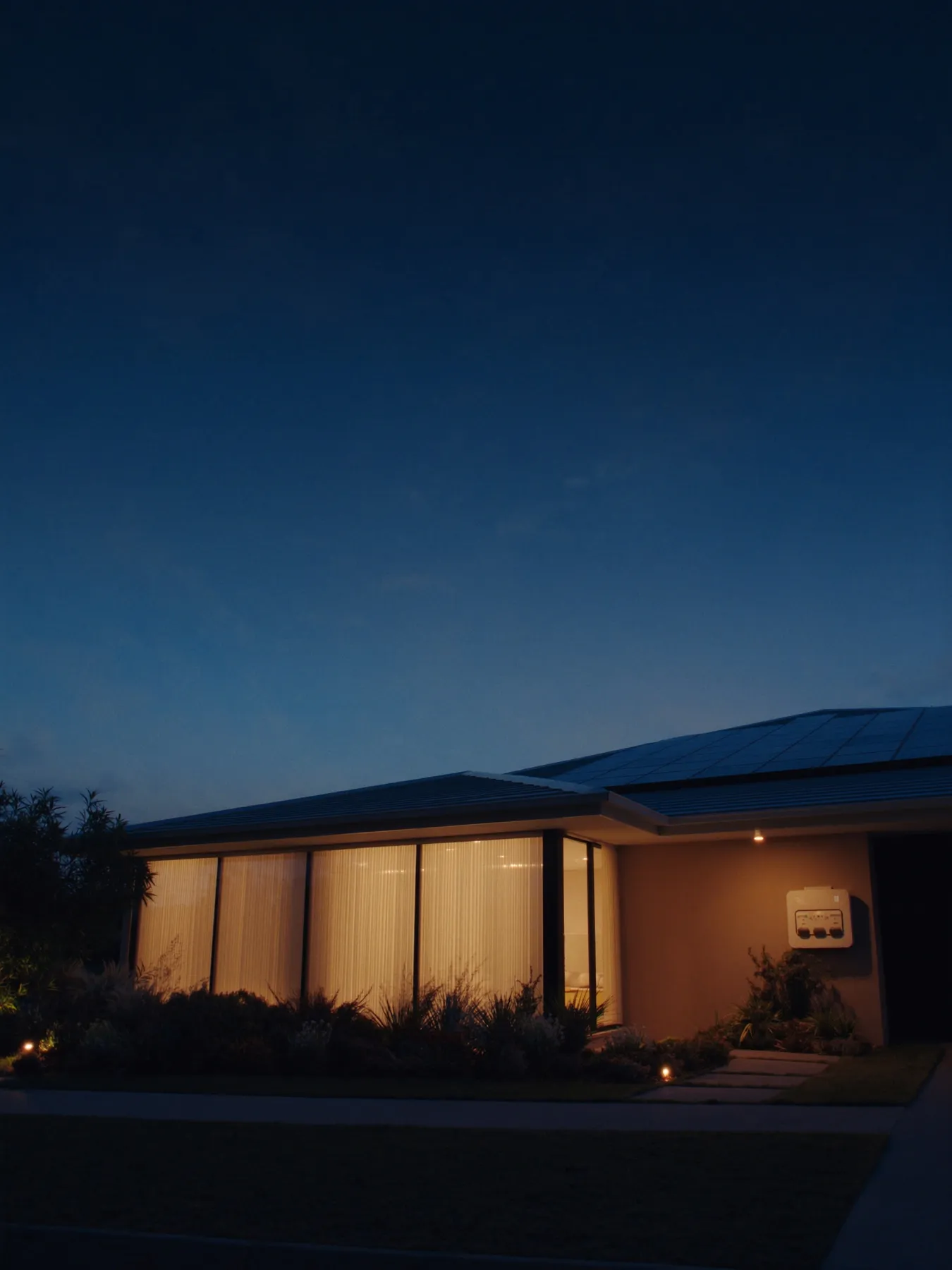 A modern Australian home glowing warm at night, running on a wall-mounted home battery